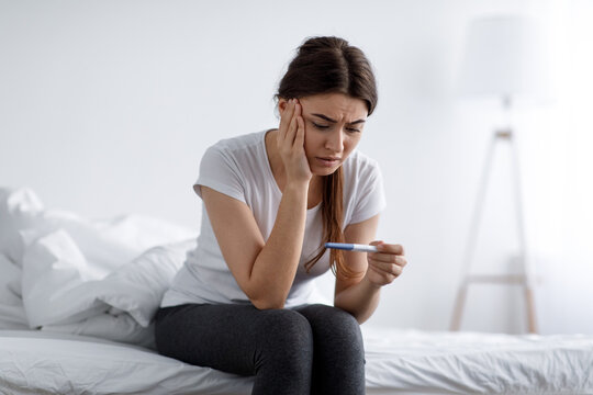 Depressed sad unhappy young pretty european woman looking at negative home test sitting on bed in bedroom interior alone