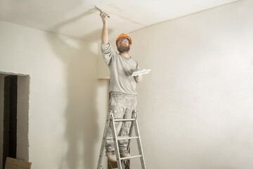 a man plasterer builder stands in a helmet and glasses with a spatula on the stairs in the room and plasters the ceiling