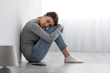 Bored upset depressed lady sitting on floor with smartphone, looking indifferent to side having...