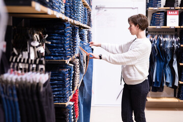 Photo of young attractive brunette woman with a short haircut in a white sweater chooses stylish and casual jeans and denim clothes in a store in a shopping centre