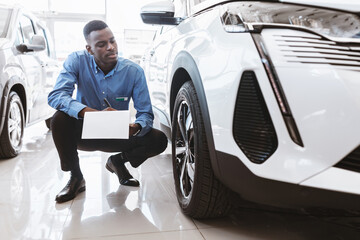 Serious black auto salesman sitting near new car, taking notes in clipboard at automobile dealership