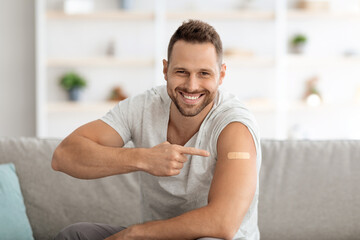 Young vaccinated guy pointing at shoulder with medical plaster and widely smiling, feeling healthy after immunization