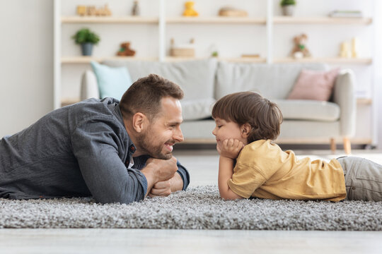 Side view portrait of handsome father and his cute little son looking at each other and smiling, lying on floor at home