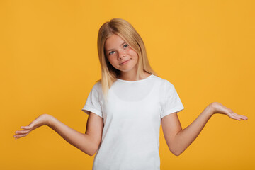 Calm confused pretty european teen girl in white t-shirt spreads her arms to the sides, isolated on yellow background