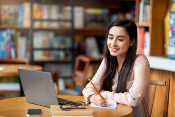 Fototapeta premium Remote education. Happy latin female student using laptop and taking notes, sitting at cafe interior, copy space