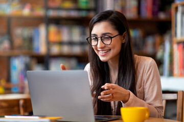 Positive arab woman having video conference on laptop, sitting at city cafe, talking and gesturing to computer webcamera