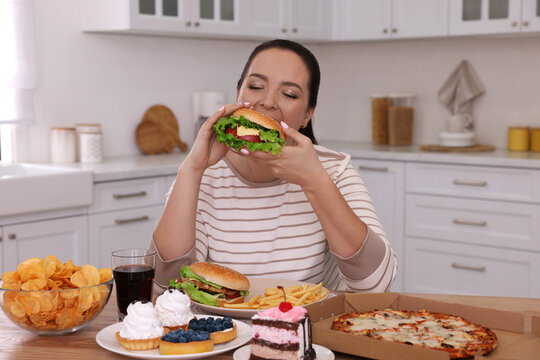 Overweight woman eating burger in kitchen at home