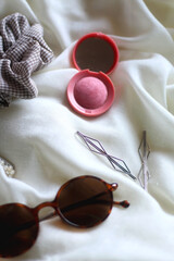 Tortoiseshell sunglasses, scrunchie, blush and various silver jewelry on white fabric background. Selective focus.