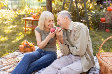 Positive senior couple sitting on field grass and enjoy eating apple, chatting and laughing, resting outdoors in garden