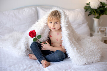 Obraz premium Beautiful blond toddler child, boy with lipstick kisses on his body, holding red rose for Valentine