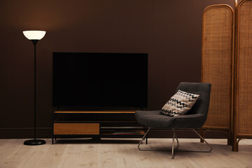 Modern TV on cabinet, armchair and floor lamp near brown wall in room. Interior design
