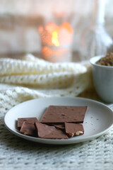 Plate with almond chocolate bar, mug with chocolate chip cookies, open book, reading glasses, soft blanket and lit candles. Hygge at home. Selective focus.