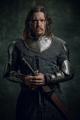 Half-length portrait of brutal seriuos man in image of medieval warrior or knight with dirty wounded face holding big sword isolated over dark background.