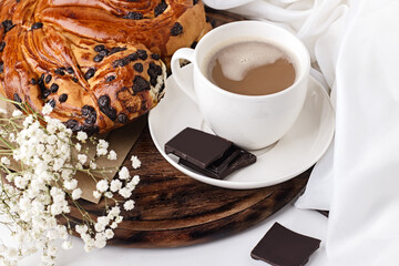 A cup of coffee with milk and a fresh bun with pieces of chocolate on a light background.