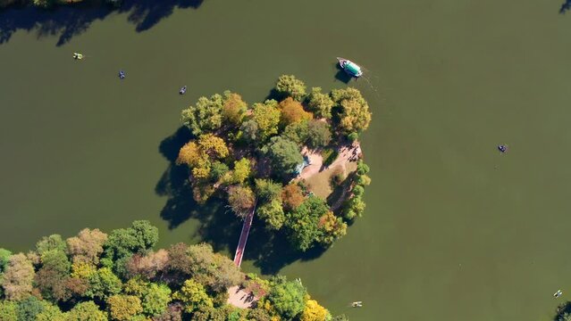 Top view of the lake and the island. People swim on a boat, boats on the lake. Yellow, green, autumn leaves, trees from above. People are walking in the park.