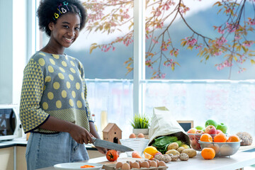 Dark skinned teen girl preparing fresh salad in kitchen at home, cutting tomato, near the window...