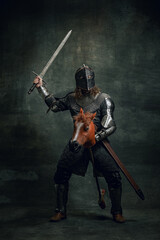Portrait of medieval warrior or knight wearing helmet and armor riding toy horse, holding big sword isolated over dark background. Comparison of eras, history