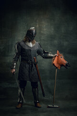 Portrait of medieval warrior or knight wearing helmet and armor riding toy horse, holding big sword...