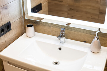 Modern washbasin with chrome faucet beside a stylish soap dispenser. Mirror with built-in led...
