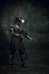 Portrait of man, medieval warrior, knight with dirty wounded face in VR headset isolated over dark vintage background. Comparison of eras, history