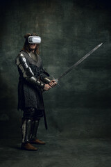Fototapeta premium Portrait of man, medieval warrior, knight with dirty wounded face in VR headset isolated over dark vintage background. Comparison of eras, history