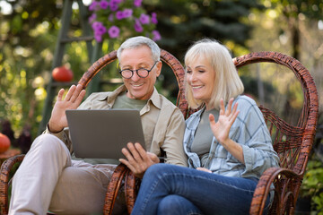 Modern grandparents. Happy senior spouses having video call via laptop, relaxing in wicker chairs in garden