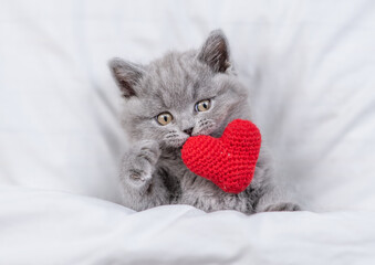 Cute kitten hugs red heart on a bed under warm white blanket. Valentines day concept. Top down view