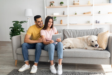 Beautiful young diverse couple sitting on couch with their cute dog, using tablet pc, browsing web at home