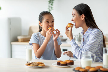 Having a bite. Cute asian girl and her mother eating snacks in kitchen, enjoying homemade muffins...