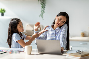 Upset asian teen girl sitting by working momin kitchen interior, asking for attention. Busy woman...
