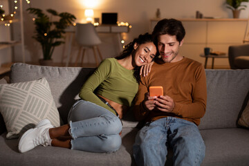 Young Couple Using Phone Browsing Internet Relaxing Sitting At Home