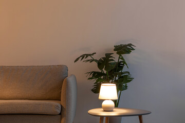 Modern sofa, glowing lamp on table in evening, plant in pot on floor on gray wall background in living room