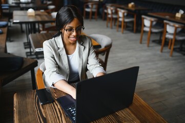 Beautiful african business woman waiting for the meeting in a co-working space and sitting with the portable computer. Hipster girl is surfing the web on a laptop during lunch.