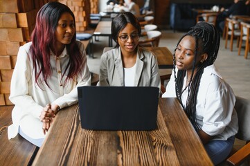 Three African American girls (students) sitting at the table in cafe studying up for test or making homework together, they are using laptop and digital tablet.