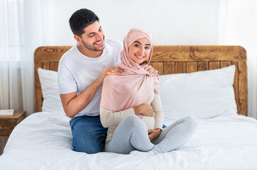 Obraz premium Caring arab husband massaging shoulders of his young arab pregnant wife, woman smiling to camera, sitting on bed