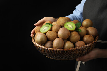 Woman holding wicker bowl with fresh ripe kiwis on black background, closeup
