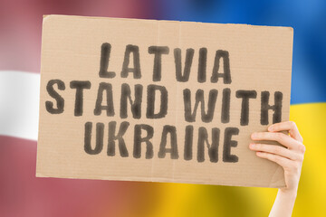 The phrase " Latvia stand with Ukraine " on a banner in men's hand with blurred Latvian and Ukrainian flag on the background. Team. European. War. Assistance. Negotiation. Safety. Security