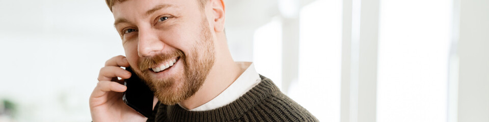 Obraz premium Young ginger man drinking coffee while talking on mobile phone in office