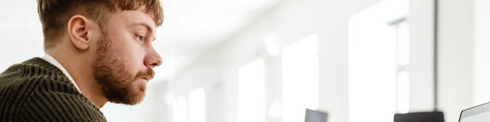 Obraz premium Young ginger man working with laptop while sitting at table in office