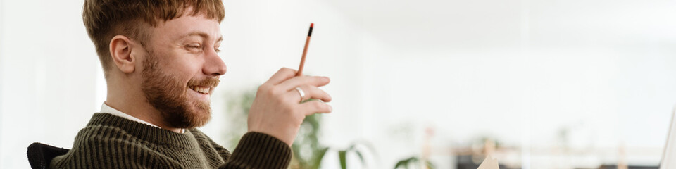 Young ginger man gesturing and working with laptop while in office