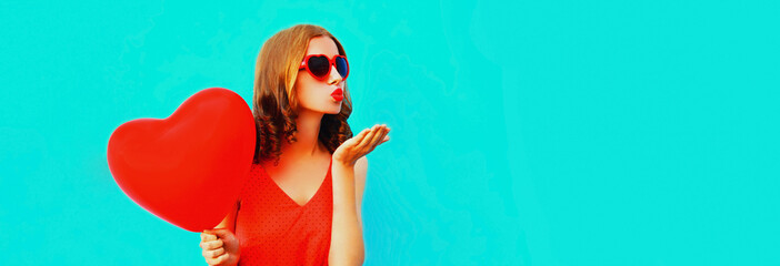 Portrait of beautiful young woman with red heart shaped balloon blowing her lips sending sweet air kiss wearing sunglasses on blue background