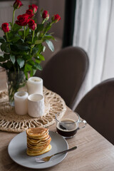 A stack of pancakes in powdered sugar with a cup of black coffee on a wooden table decorated with red roses and candles. Fresh homemade pancakes made with love for Valentine's Day