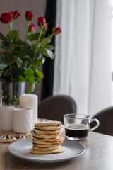 A stack of pancakes in powdered sugar with a cup of black coffee on a wooden table decorated with red roses and candles. Fresh homemade pancakes made with love for Valentine's Day