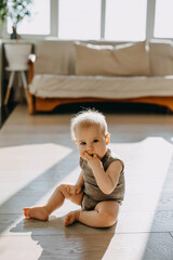 Barefoot baby sitting on a wooden floor at home, sucking fingers, looking at camera.