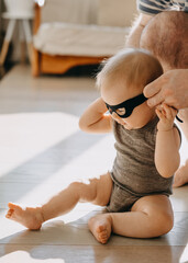 Baby sitting on the floor, wearing a superhero black mask. Father putting on baby superhero mask.