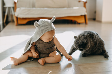 Baby sitting on the floor playing with a cat, wearing a hooded cardigan with ears.