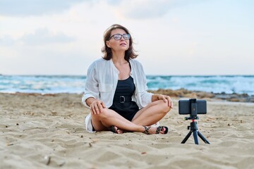 Fototapeta na wymiar Middle aged woman sitting on beach with smartphone using video call