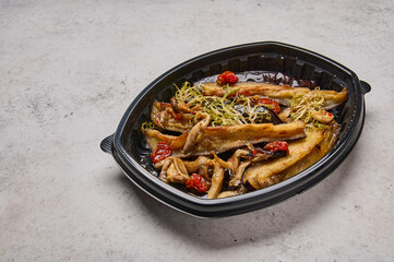 Crispy eggplant with baked cherry tomatoes and oyster mushrooms in black plastic container on light background. Designer food concept for take away or delivery