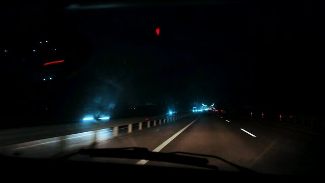 Close-up, interior of moving car along night highway, view through windshield. Road in blur, bokeh from headlights of oncoming cars, lanterns on track. Auto driving on freeway with high speed evening