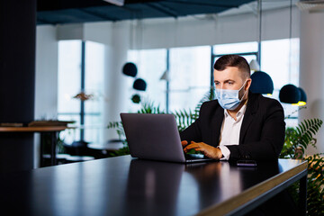 Handsome young man in a mask working in a cafe with a laptop during the coronavirus pandemic.